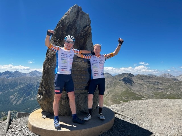 Two men onto of a mountain next to a rock in cycling clothes